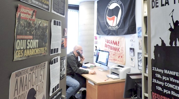 A middle-aged man in a dark button-down shirt and jeans sits at a wooden desk, looking at a white desktop iMac computer while using a black landline phone. He is sitting by a window through which light is shining. To his left and right are anarchist posters decorating the doorway of his office.