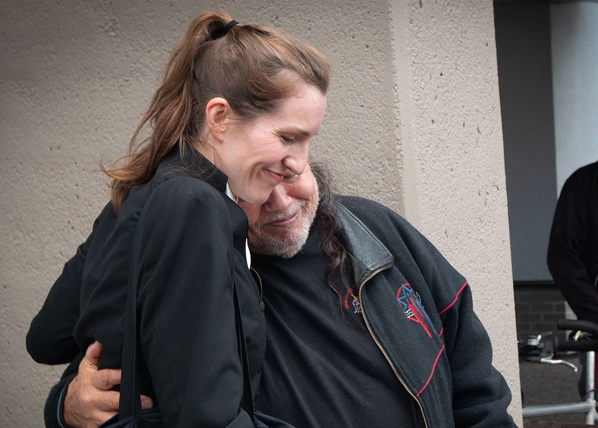 Two people hug. One is a smiling light-skinned woman in lawyer&rsquo;s black robes, the other a middle-aged man wearing black, with. greying beard and gentle smile. 
