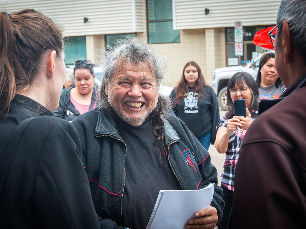 A middle-aged man with medium-toned skin grins at another person. He has somewhat messy grey hair and stubble, and wears black. He is outside a courthouse.