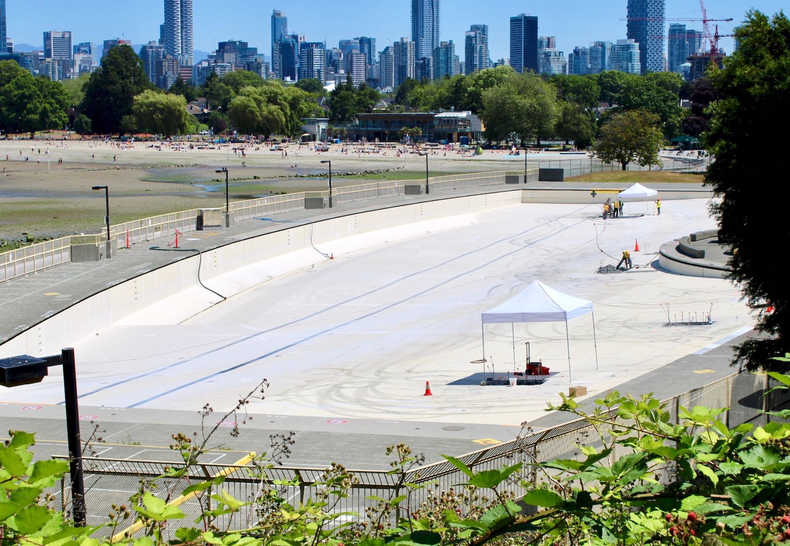 An outdoor pool sits empty, with a handful of construction workers visible within it. The downtown Vancouver skyline can be seen in the background.
