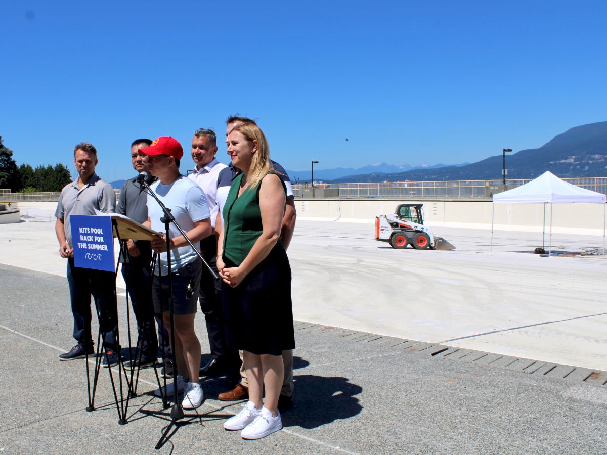 Six people stand behind a portable podium giving a press conference inside the empty Kitsilano Pool. A blue sign on the podium says 'Kits Pool Back for the Summer.'