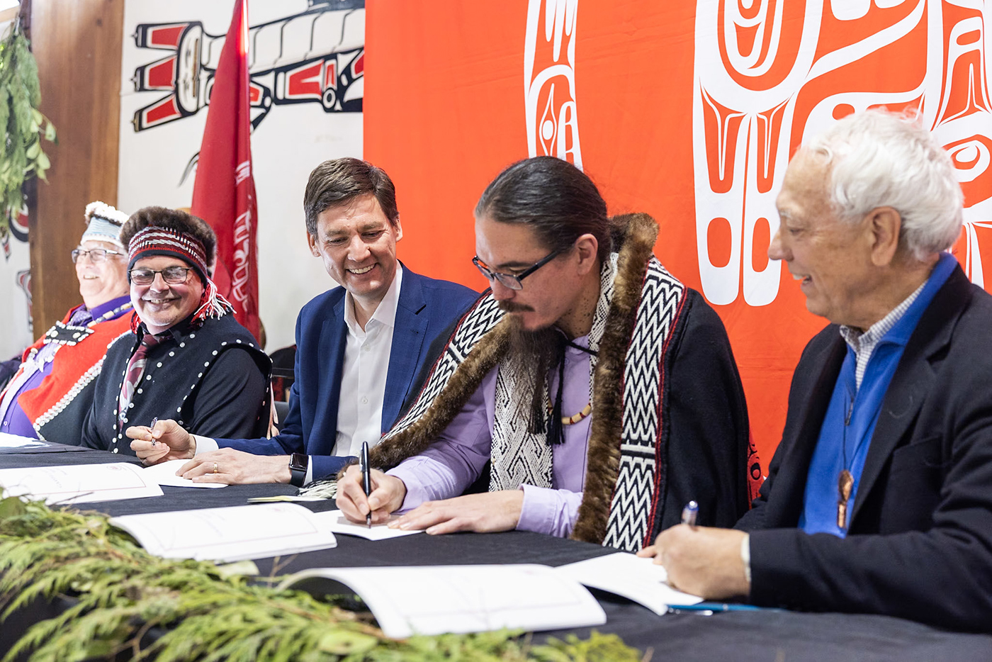 Five people sit in a row at a table. Near the centre, a man with a ponytail and goatee beard, wearing glasses and traditional Indigenous regalia, holds a pen as he signs a piece of paper.