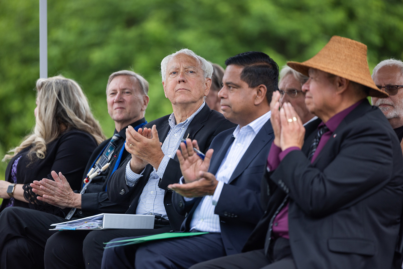 Several people sit in a row of chairs in an outdoor setting clapping their hands.