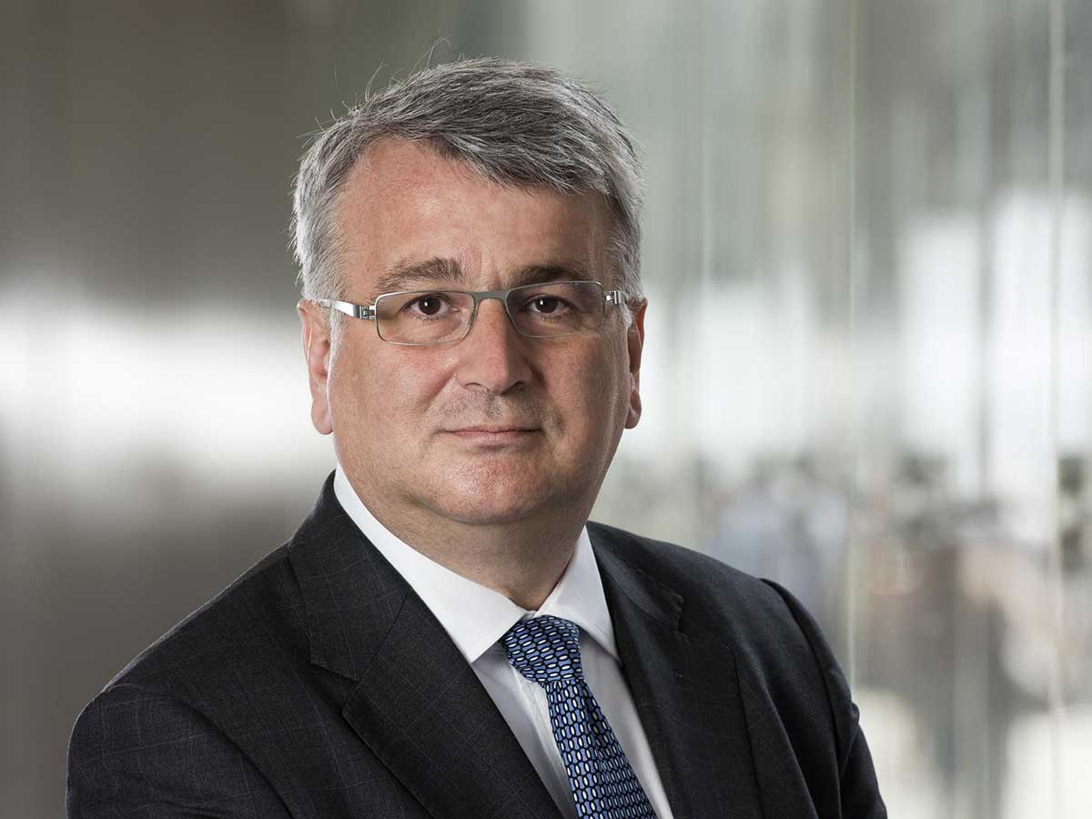 A lighter skinned man with grey hair and grey framed glasses looks into the camera. He is wearing a dark suit, a white shirt and a patterned tie.