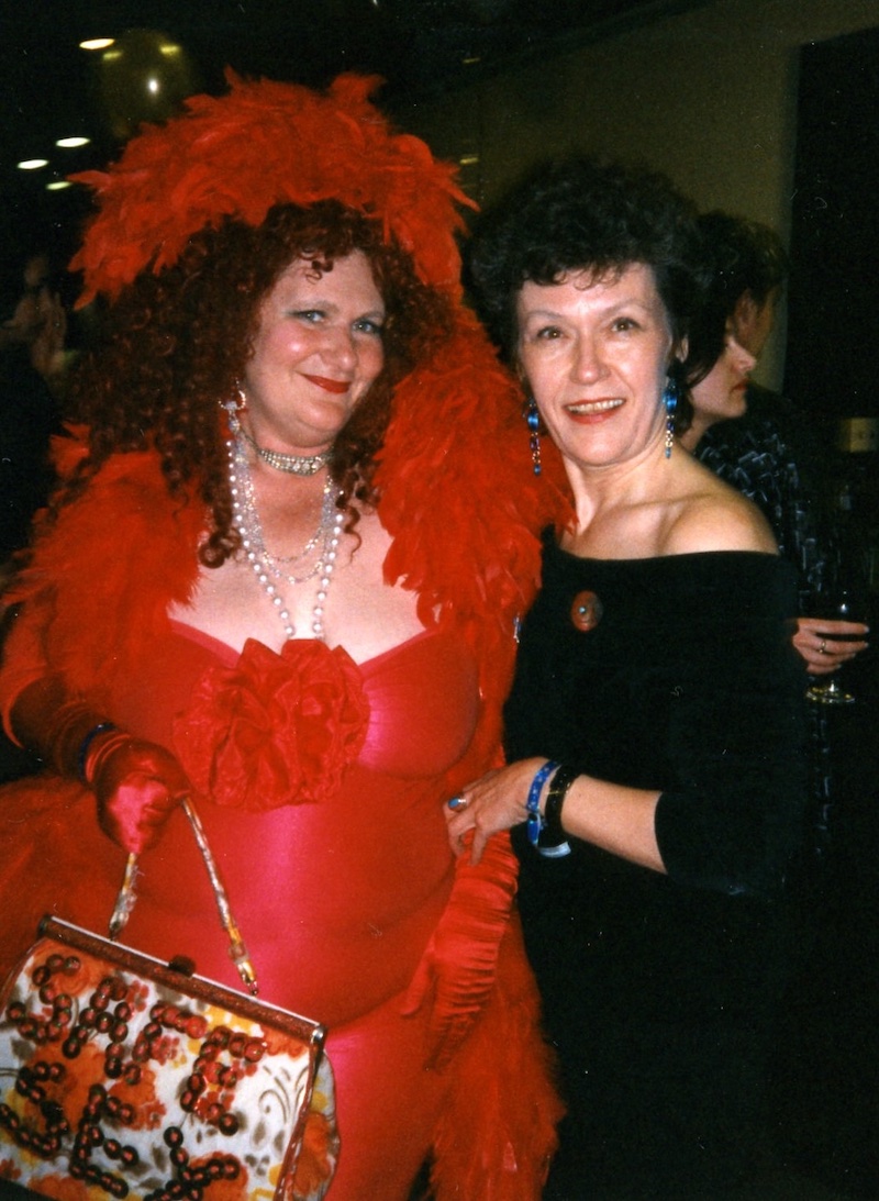 Two people pose in a party photo from 1997. To the left, Carol Leigh stands in a red dress and feather boa. To the right, Frances Shaver wears a black dress that reveals her shoulders.
