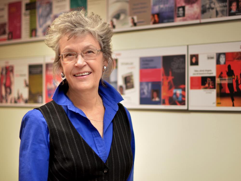 Frances Shaver has short wavy grey hair, glasses and light skin. She is smiling and standing against a wall mounted with rows of framed publications. She wears an indigo button-down under a black vest.