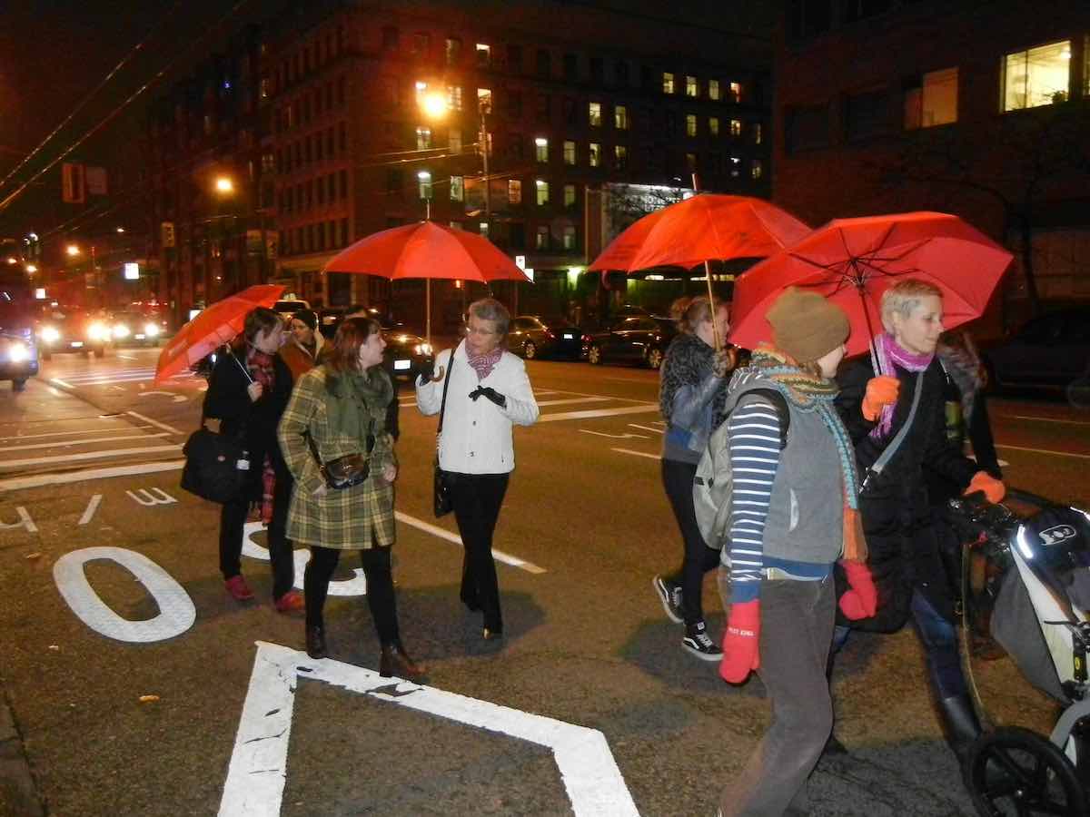 People holding red umbrellas walk down a downtown street at night wearing winter clothes. Frances Shaver, centre, wears a white jacket and holds a red umbrella while speaking to someone in a plaid coat.