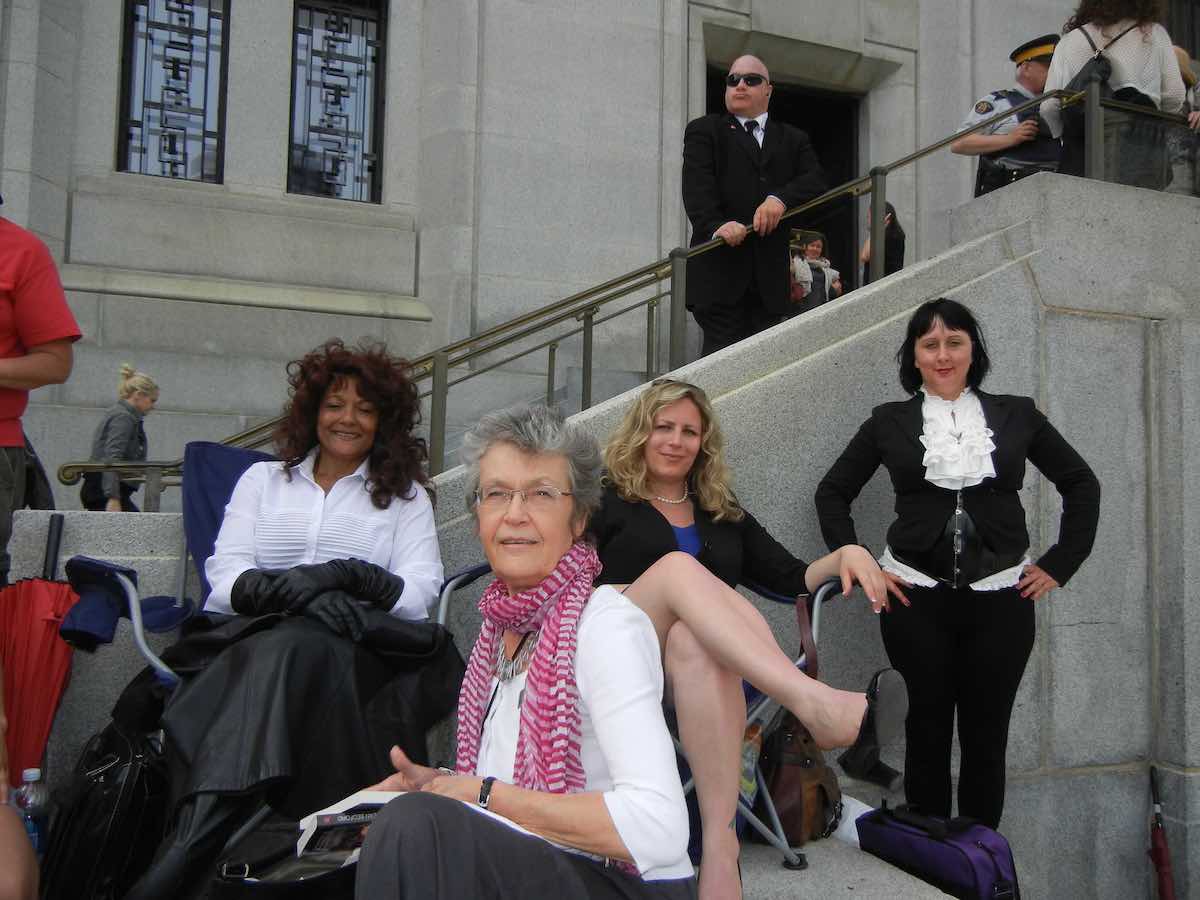 Four women pose together against a concrete staircase in front of the Supreme Court of Canada. Frances Shaver is in a white shirt and red scarf at centre; Terri-Jean Bedford is in a white shirt and holds a black jacket to the left of Shaver.