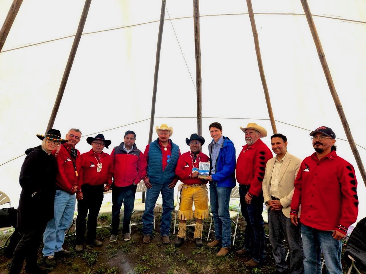 A group of people, many wearing red shirts and cowboy hats, stand in a semicircle inside a tent-like structure. At the centre, two men jointly hold a book between them.