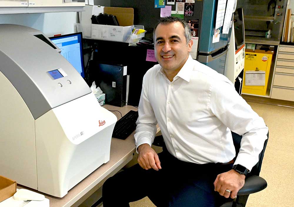 A middle-aged man with salt and pepper hair is wearing a white collared shirt and black pants. He is sitting in front of a computer screen partially hidden by a large, bulky grey and cream-coloured machine. He is turning towards the camera and smiling. Behind him several other lab machines are visible, including a teal-coloured fridge.