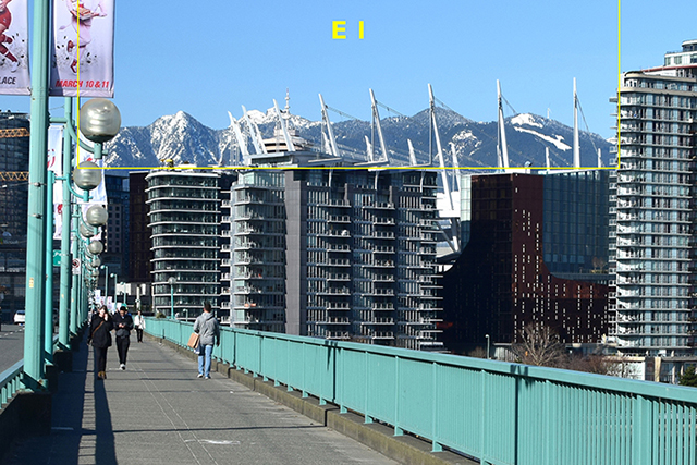 A view of downtown Vancouver from the Cambie Bridge. In the foreground is a pedestrian walkway with turquoise railings on the right. In the background are the snow-capped North Shore mountains visible across rows of condo towers. Sharp white spires poke up among them. They are outlined in yellow and labelled 'E 1.'