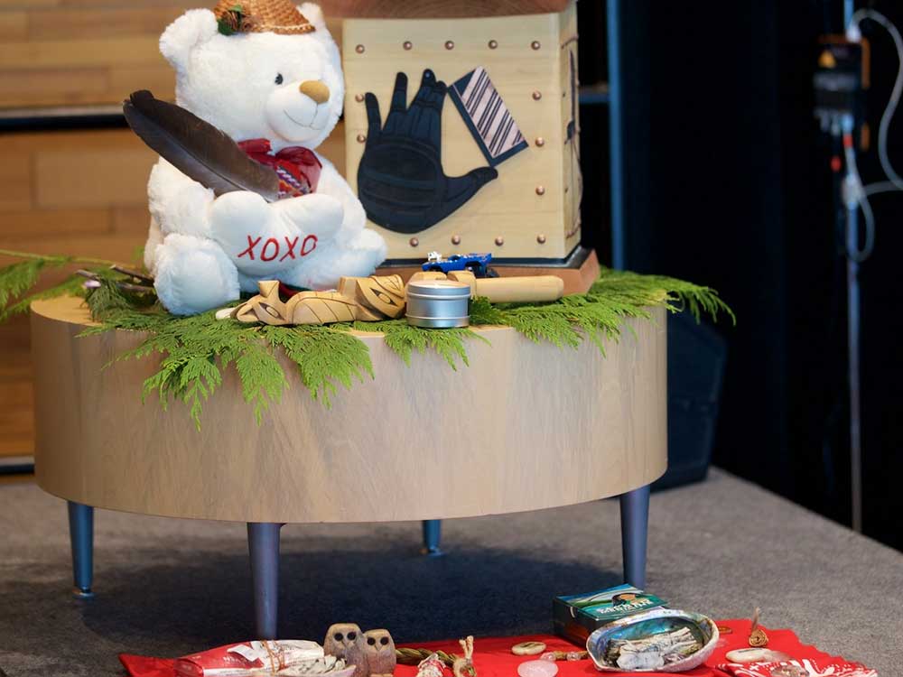 A white teddy bear wears a miniature woven cedar hat and sits on fresh cedar bows on a small, low, light birch table. Around it are memorial items including Indigenous carvings, artwork and offerings such as sage, a red blanket, stones and feathers.