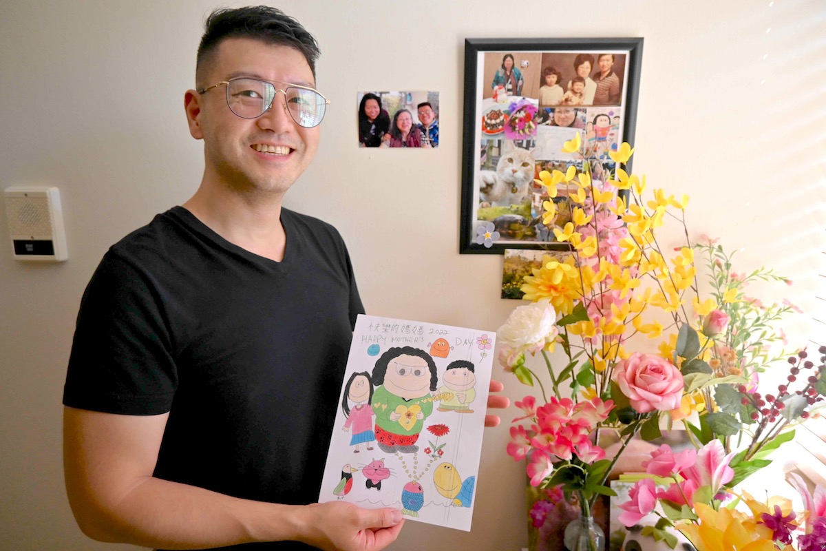 Eric Li stands before a wall of framed photos and a bright fake floral arrangement. He is a Chinese man in black summer clothing and glasses. He holds a piece of artwork that he and his mother made together.