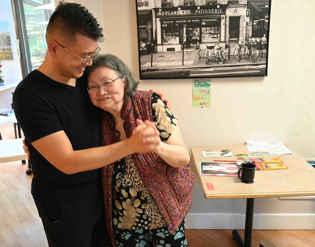 Eric Li, left, stands with one arm around his mother, May Tsoi, right, looking down at her and smiling. His other hand is clasped with hers as she leans against his chest, smiling. Li is wearing black summer clothing and his mother is in a colourful red vest over animal print clothing.
