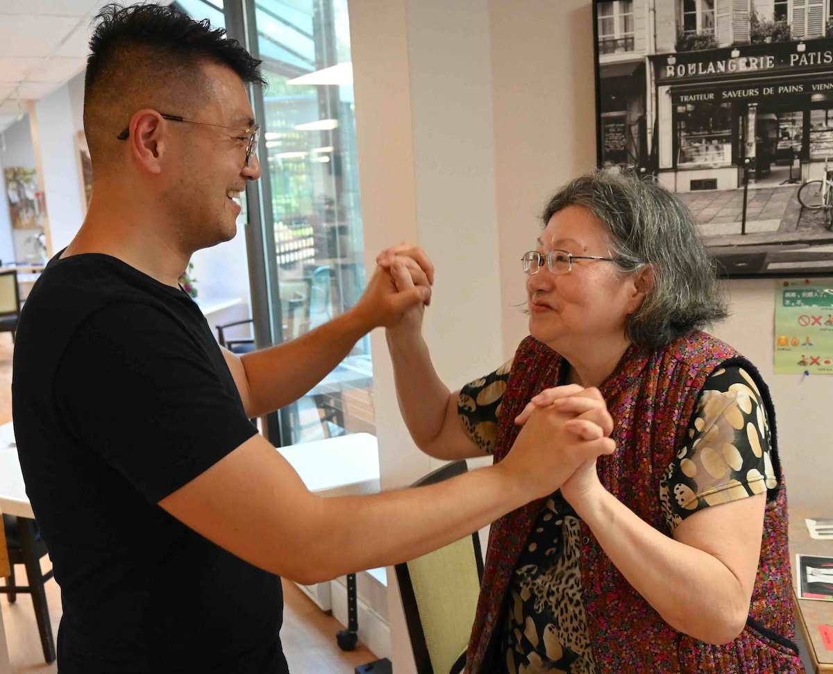 Eric Li, left, clasps hands with his mother, May Tsoi, right. They are smiling at each other and dancing in a long-term care facility. Behind them is a black and white piece of art on the wall and an assortment of tables and chairs. They both have dark hair and glasses.
