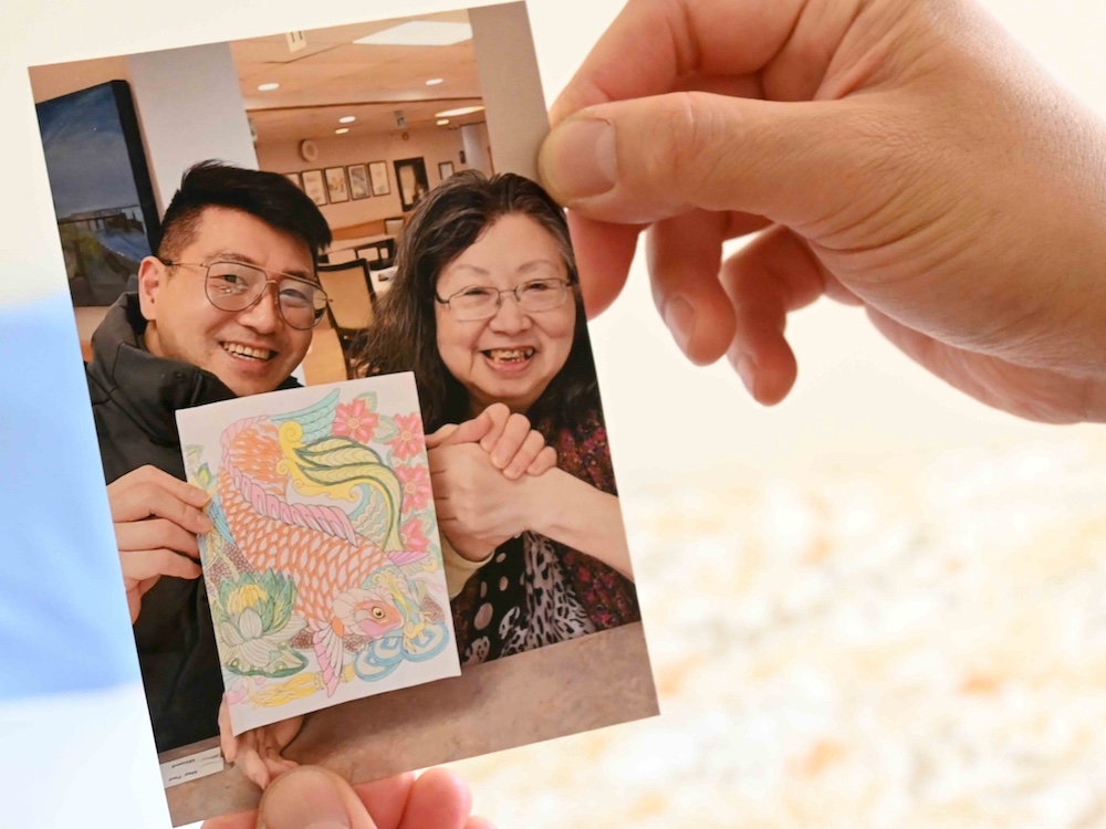 A photo of two hands holding a photograph featuring a Chinese man holding the hands of his elderly mother in an indoor space, smiling. They are both wearing glasses and have black hair; the man on the left holds a colourful illustration of a koi fish.