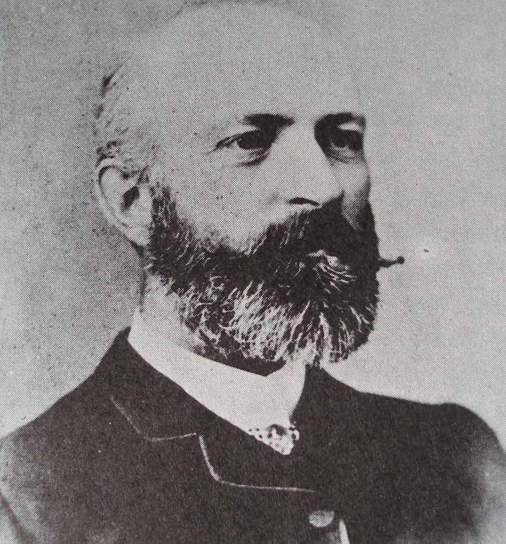 A black and white photo of a stern-looking balding man, with a bushy beard and moustache, wearing a dark suit.