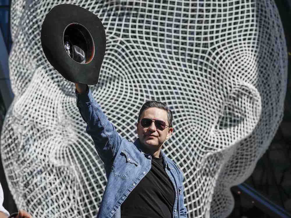 Pierre Poilievre has short, slicked-back, dark brown hair, light skin and aviator sunglasses. He is wearing a blue denim shirt over a black top and waving a black cowboy hat in the air. Behind him is a large sculpture of a human face composed of white lines in an intricate grid pattern.