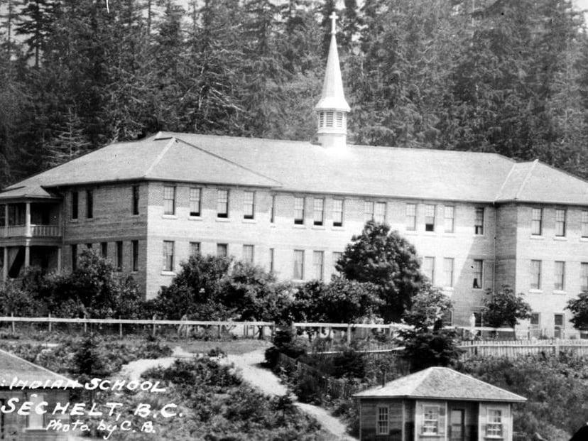 A black and white photo shows a large three-storey structure with a steeple, and forest in the background.