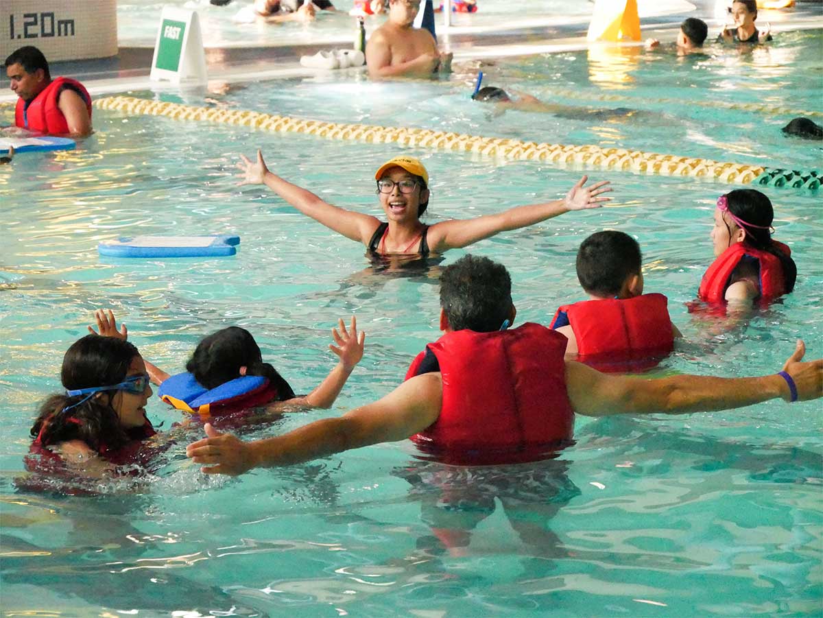 Five people wearing life jackets, four children and one adult, are floating or standing in a swimming pool, facing away from the camera. They are facing one person wearing a bathing suit, baseball cap and glasses, standing in chest-deep water with their arms spread wide.