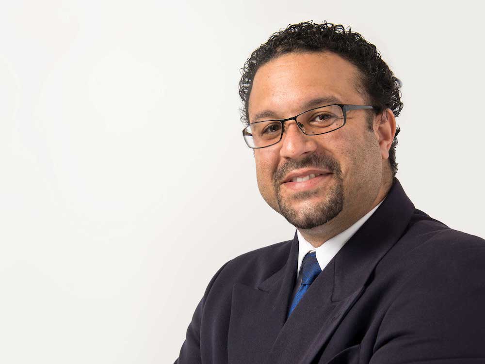 A man with short, dark curly hair and medium skin tone, wearing glasses and a black suit jacket, smiles for the camera with his arms crossed.