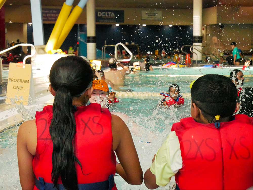 Two children, one with long dark hair in a ponytail and the other with short dark hair, are photographed from behind and are wearing red life jackets. In front of them children are splashing in an indoor pool.