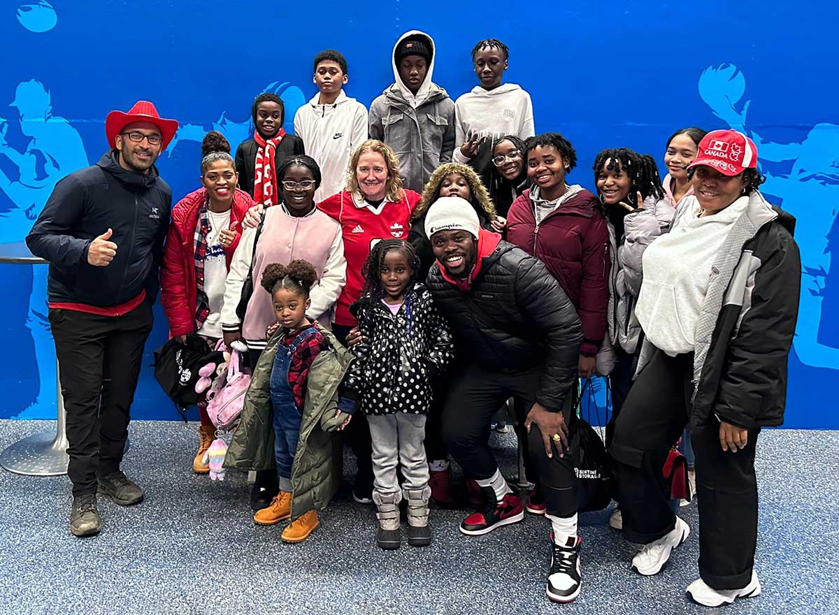 Seventeen people ranging in age from adults to young children and in skin tone from dark to medium light are standing in a huddle facing the camera. All but one of them are wearing winter jackets, and they are all smiling.