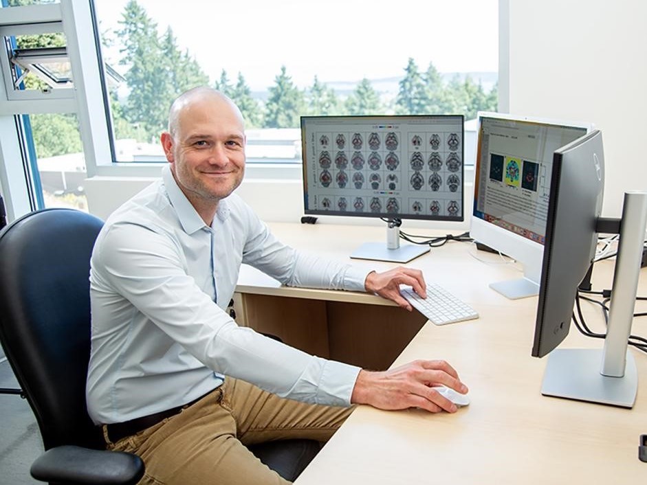 A bald white man sits at a desk with two computer screens, one showing X-rays of human brains.