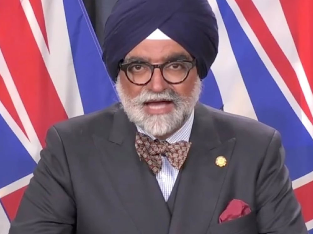 A middle-aged man with medium skin tone wearing a blue turban, suit, bow tie and tortoiseshell glasses stands at a podium with B.C. flags behind him.