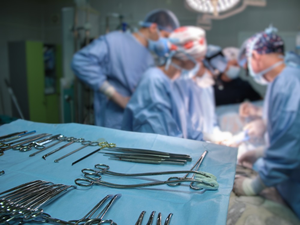 Forceps and other surgical tools sit on a tray. Three people wearing surgical garb are in the background.