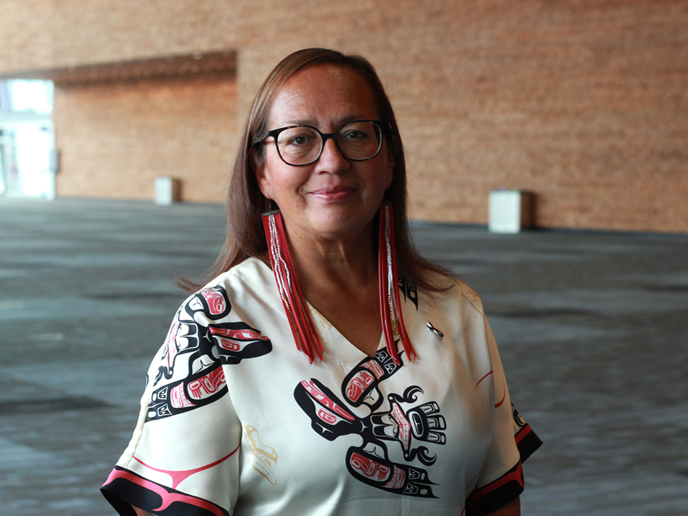 A woman with medium skin tone and long, straight brown hair looks directly at the camera. She is wearing long, beaded earrings, glasses and a white silk shift with coastal First Nations art.