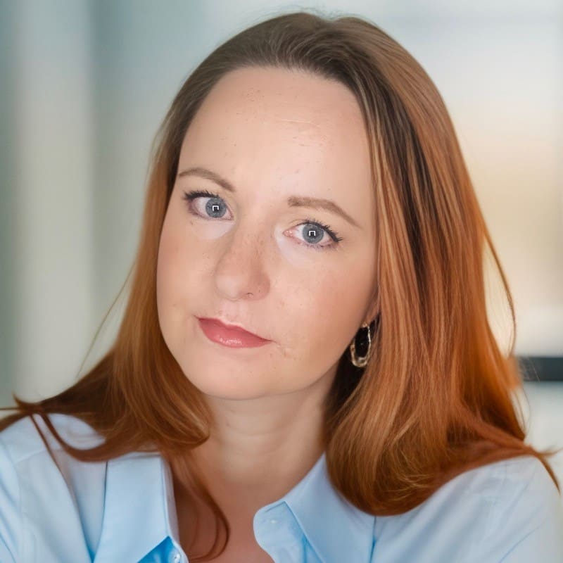 Jillianne Code, a woman with a light skin tone and long, straight red hair, looks directly at the camera. She is wearing a blue button-up shirt with the top button open.