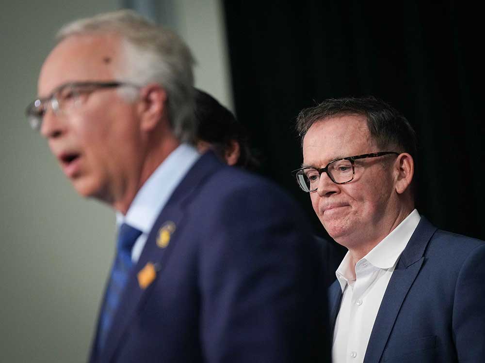 Two 60-ish men with light-toned skin, wearing blue suits, share a podium.