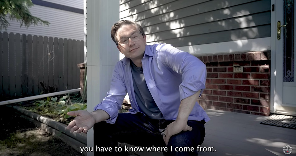 Pierre Poilievre sits on the front steps of a house wearing an unbuttoned light blue shirt over a grey-blue T-shirt. He gestures at the camera, smiling slightly. He has light skin, glasses and short dark hair.