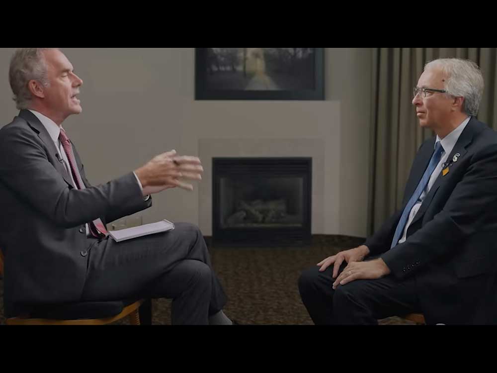 Two men in their 60s sit facing each other. They both have grey hair and wear suits and ties.