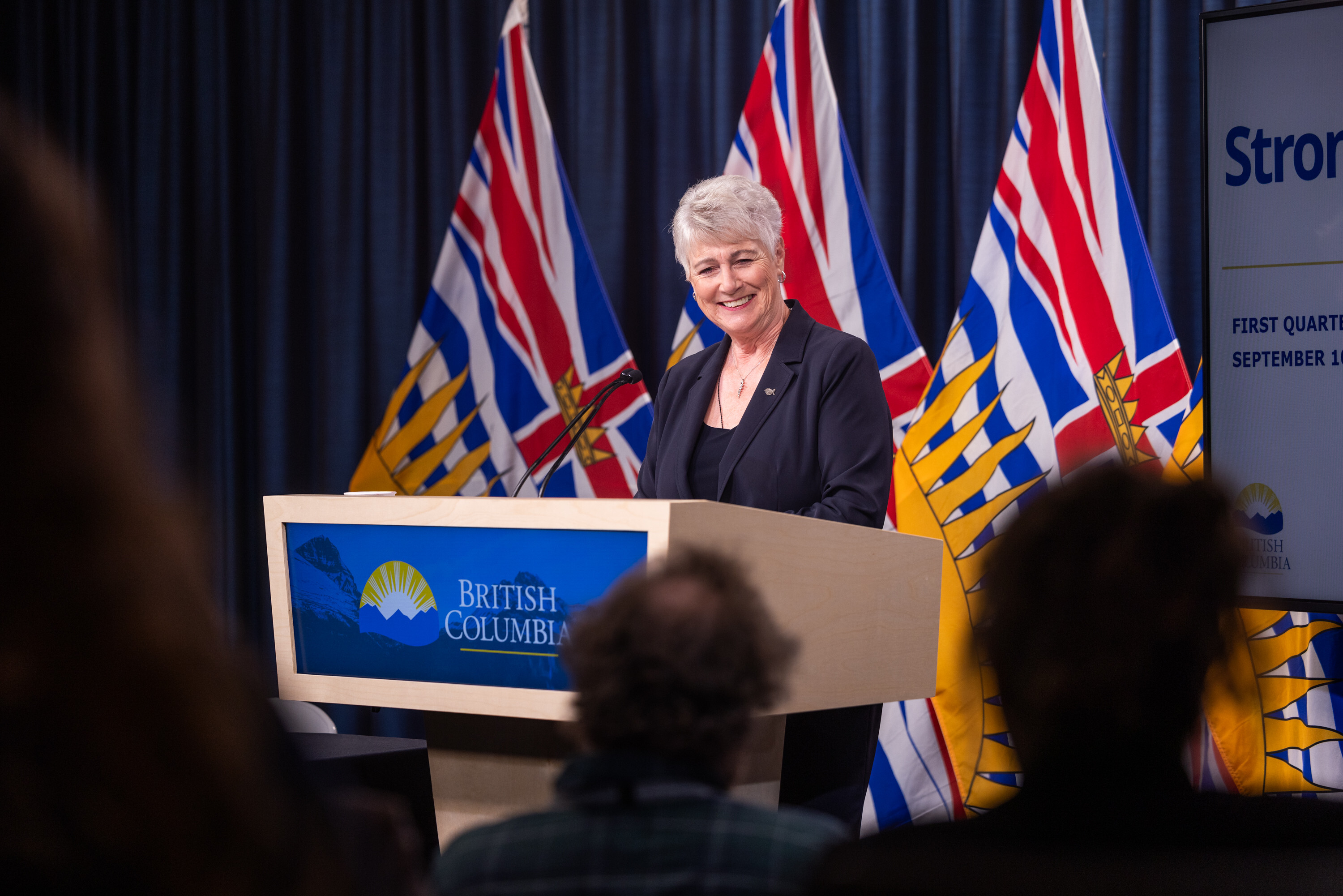 A light-skinned, grey-haired, middle-aged woman stands at a podium wearing a dark blazer and shirt.