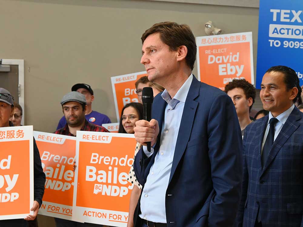 A very tall light-skinned man holds a microphone while a man with medium-toned skin and other people stand behind him. There are orange political campaign signs behind the two principal figures.
