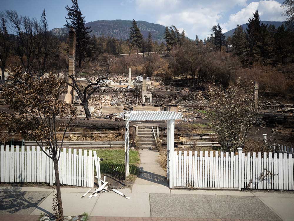 A white picket fence is mostly intact. Beyond it, many houses have burned to the ground, leaving only their charred chimney stacks.
