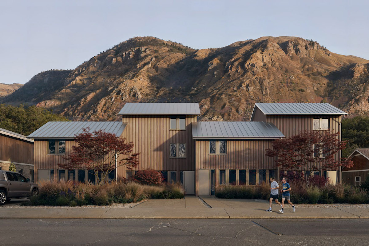 A row of beige townhouses stands against a ridge of desert hills in summer. Two men jog on the sidewalk to the right of the frame.