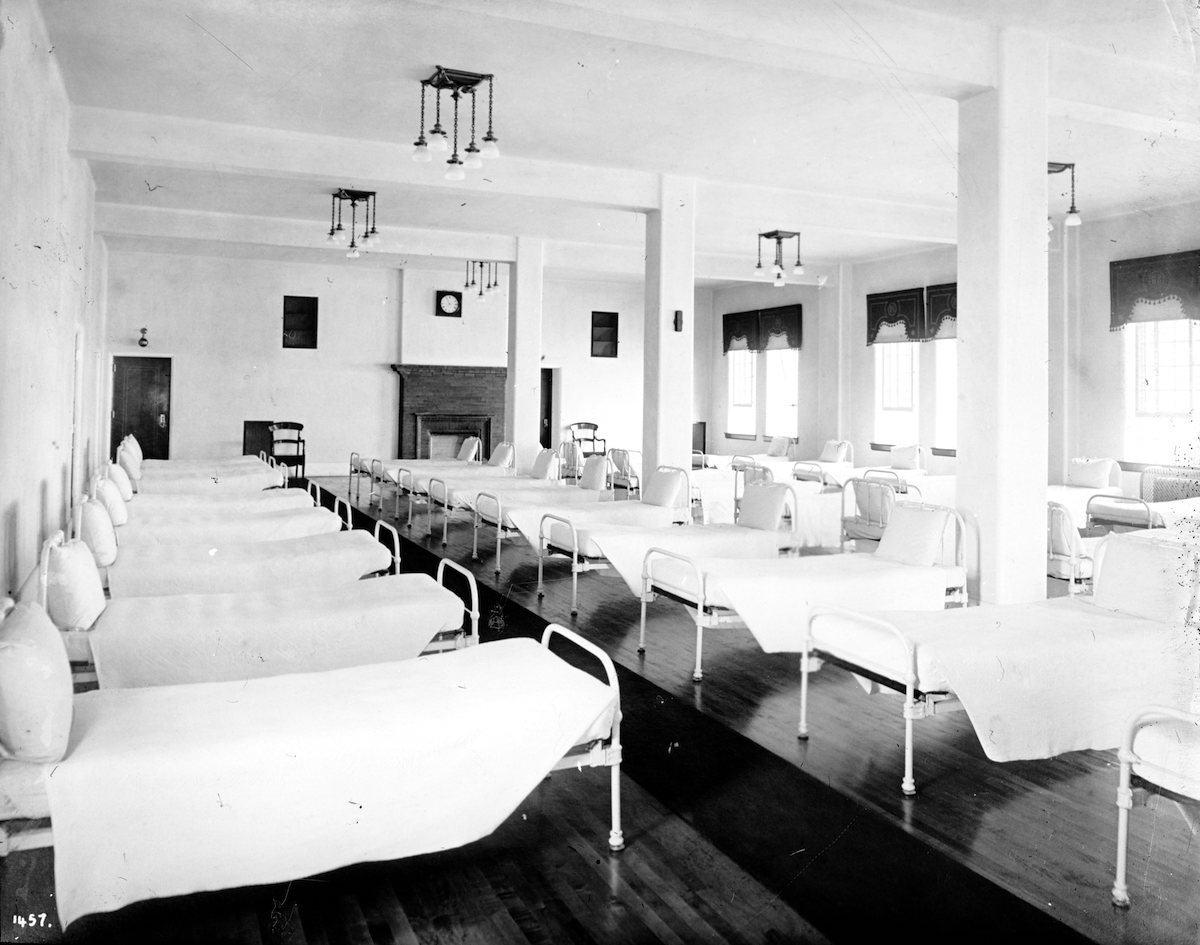 Four rows of beds are shown in a dormitory in Riverview Hospital. The beds look quite narrow and hard and institutional, and are quite close together.