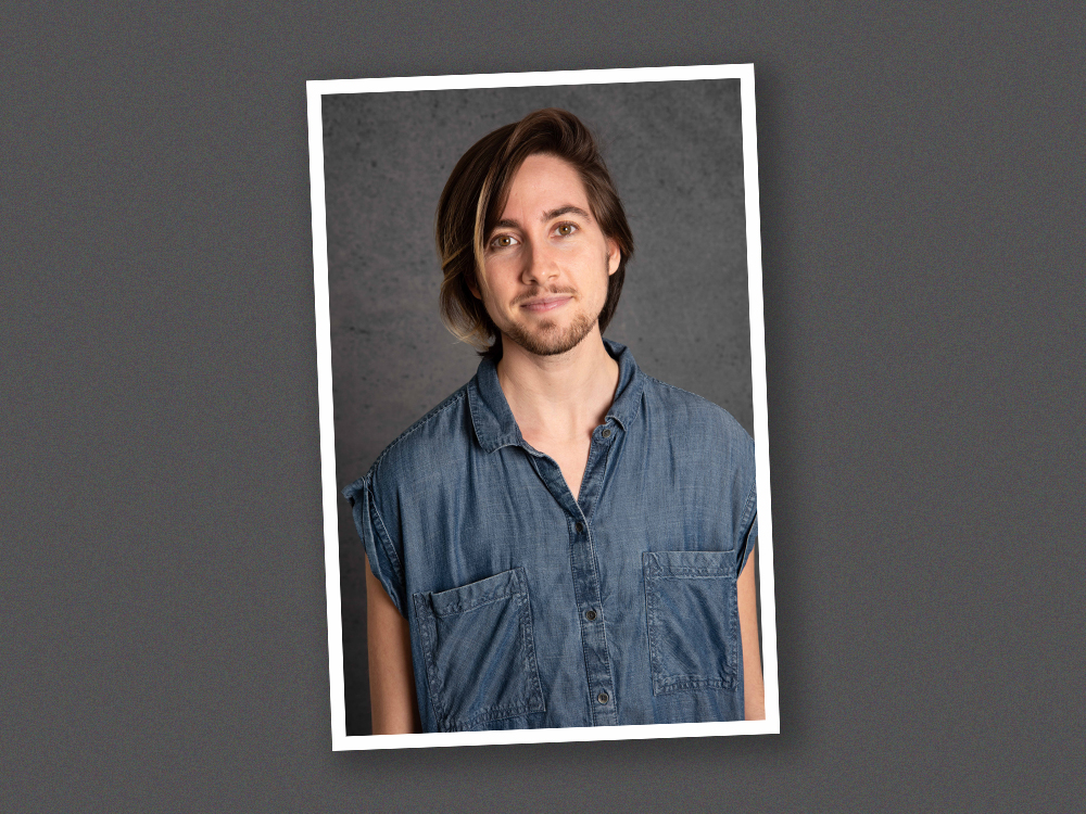 Gavin Somers has a light skin tone and shoulder-length brown hair. They have a moustache and goatee and are wearing a denim shirt.