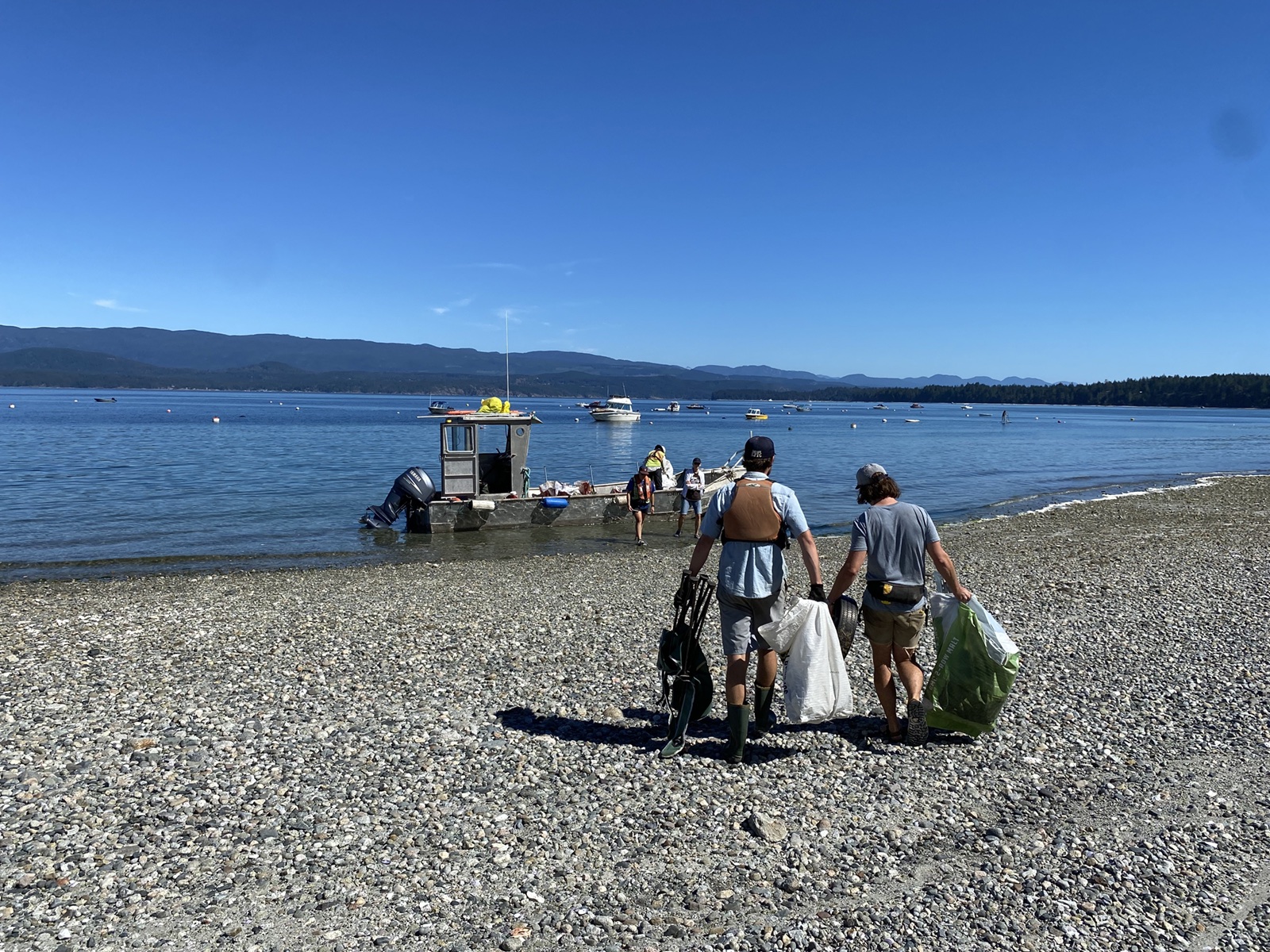 People carry ocean debris along a rocky beach towards a boat on a sunny day.