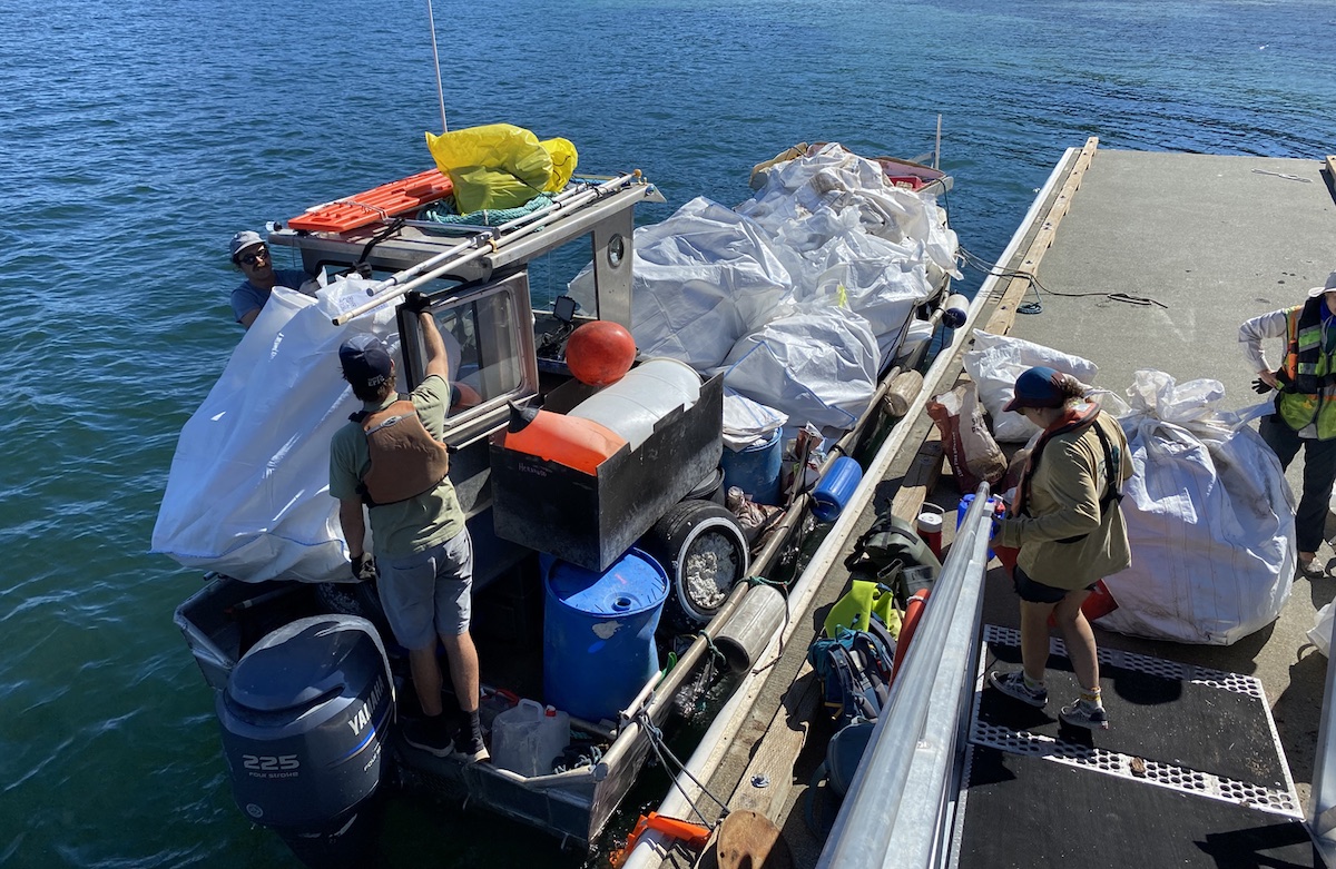 People load a small boat with large white bags of ocean debris.