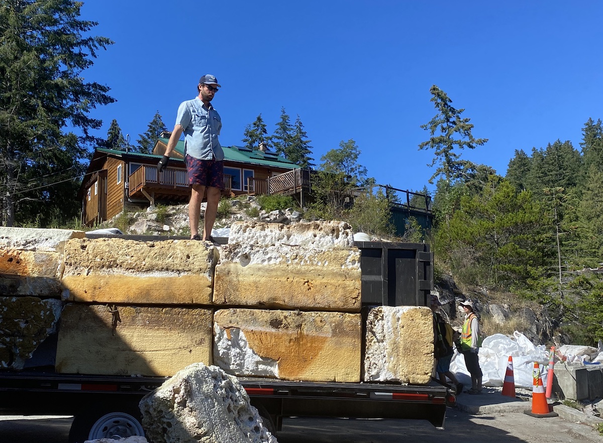 A person in a denim shirt and blue swim trunks stands atop foam blocks on a pickup truck. A large wooden house is behind him.