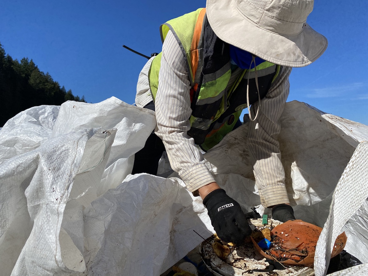 A person in a white sun hat and orange utility vest sorts debris over a large white bag.