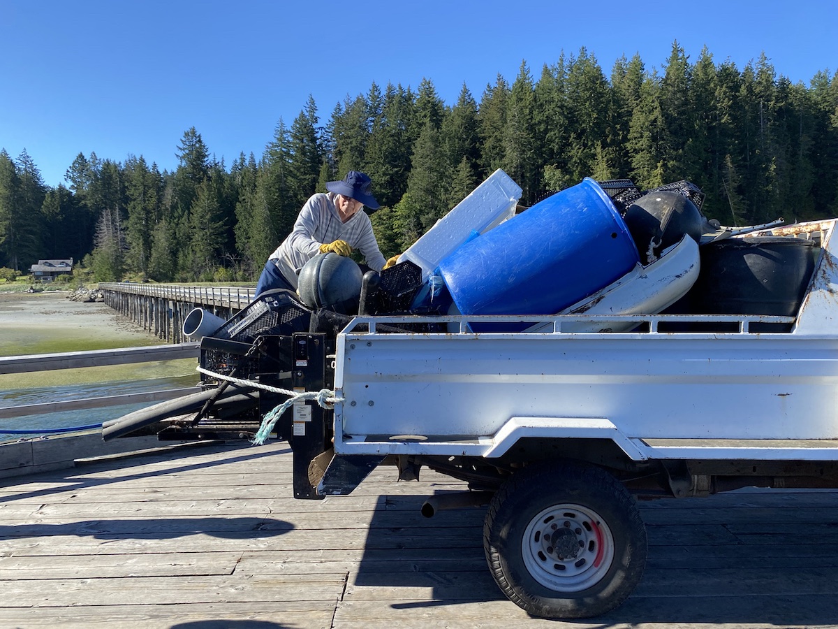 The back of a white pickup truck is loaded with ocean debris on a dock. A person in a navy sun hat and grey striped hoodie holds on to some of the material with orange gloves.