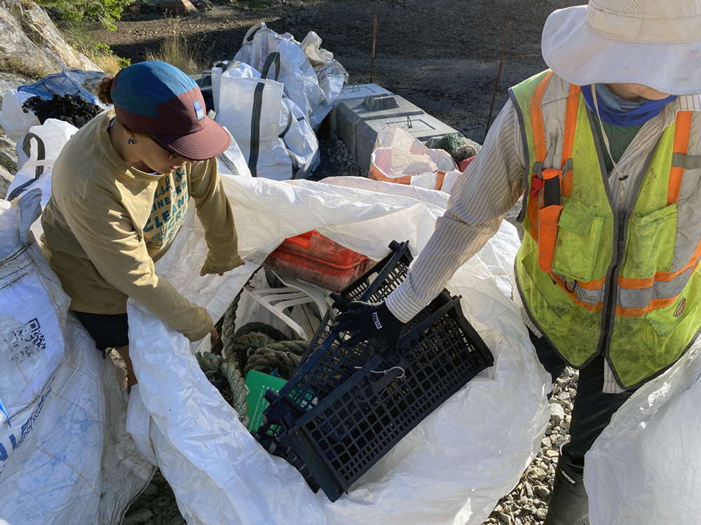Two people stand among an assortment of large white bags outdoors. On the left, a person in a mustard-yellow long-sleeved T-shirt and brown and black ball cap is holding open one bag while a person on the right in a white sun hat and high-visibility vest pulls out a black plastic basket.