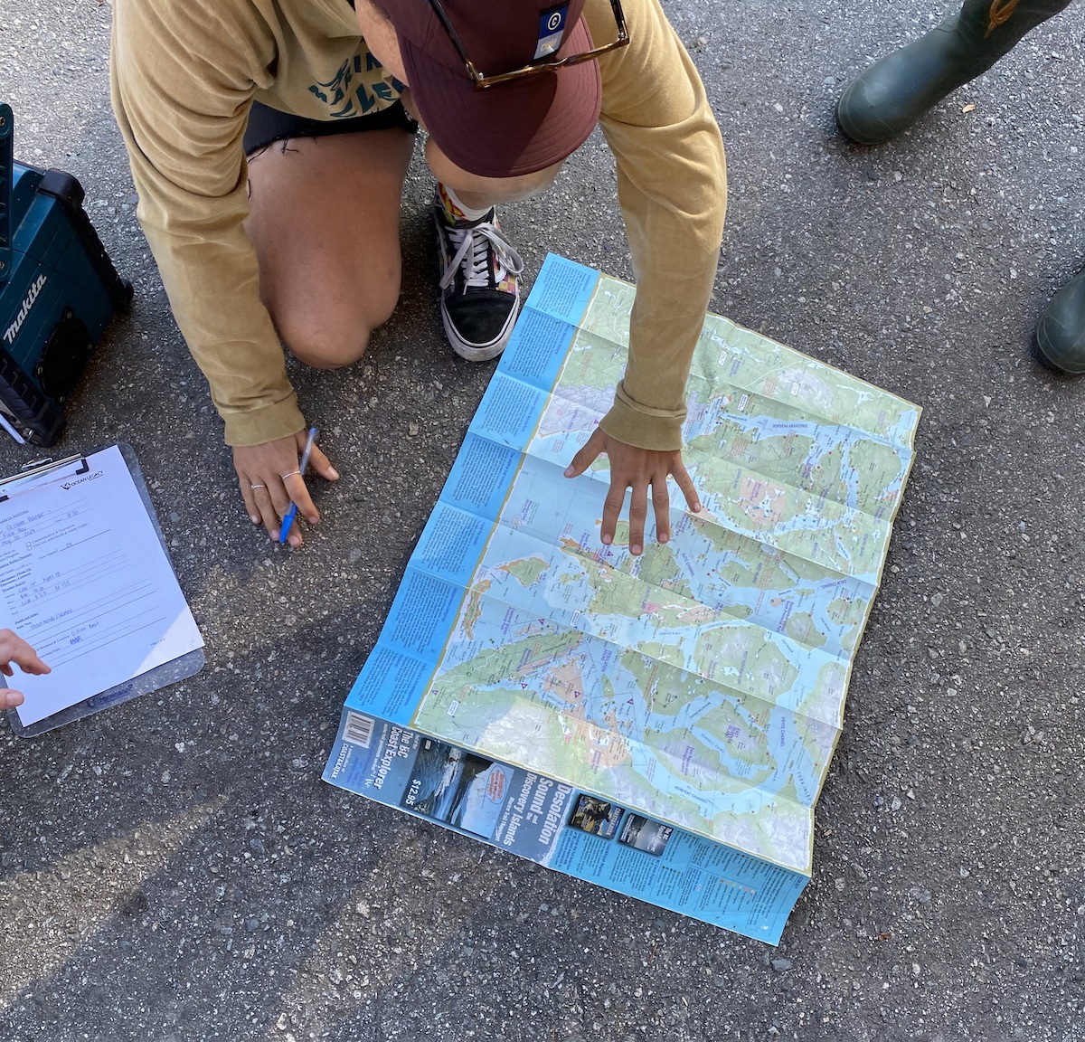 A person in a mustard-yellow long-sleeved T-shirt crouches over a blue and green map laid out on grey concrete. Another person in dark green rubber boots stands nearby.