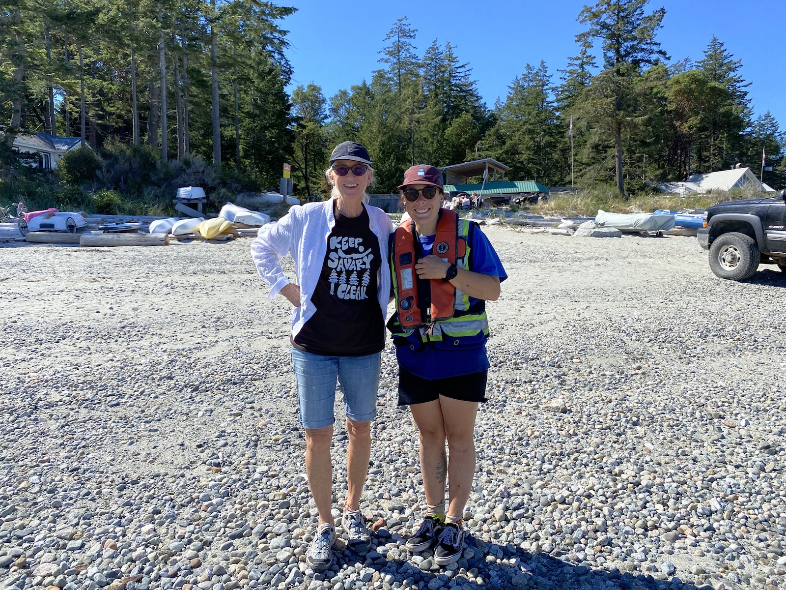 Two people stand on a rocky shoreline. On the left is a person with a black ball cap and sunglasses with a white button-down shirt over a black graphic T-shirt that says 'Keep Savary Clean.' On the left is a person with a burgundy ball cap, a utility vest and a red life jacket.