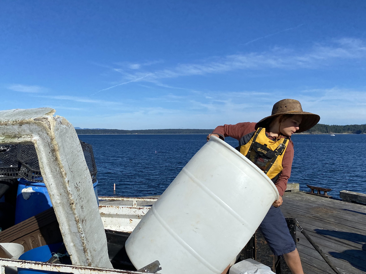 A young teen in a broad-brimmed brown sun hat hauls a light grey barrel on a ship on the ocean on a bright day against a blue sky.